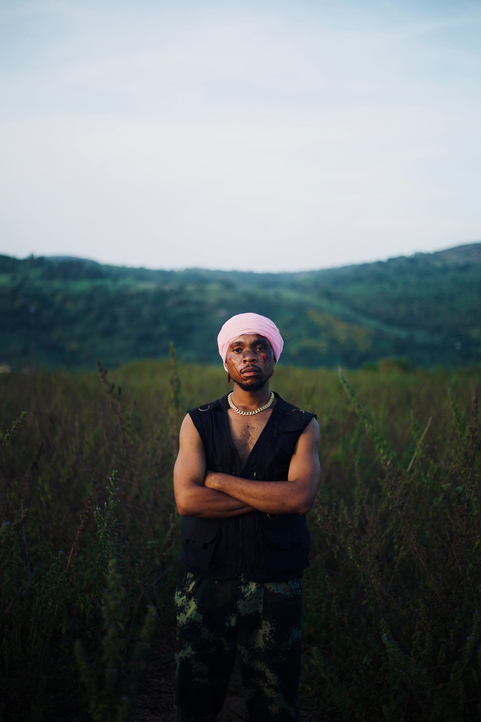 An artist in a pink headwrap stands with arms crossed in tall grass, green hills behind.