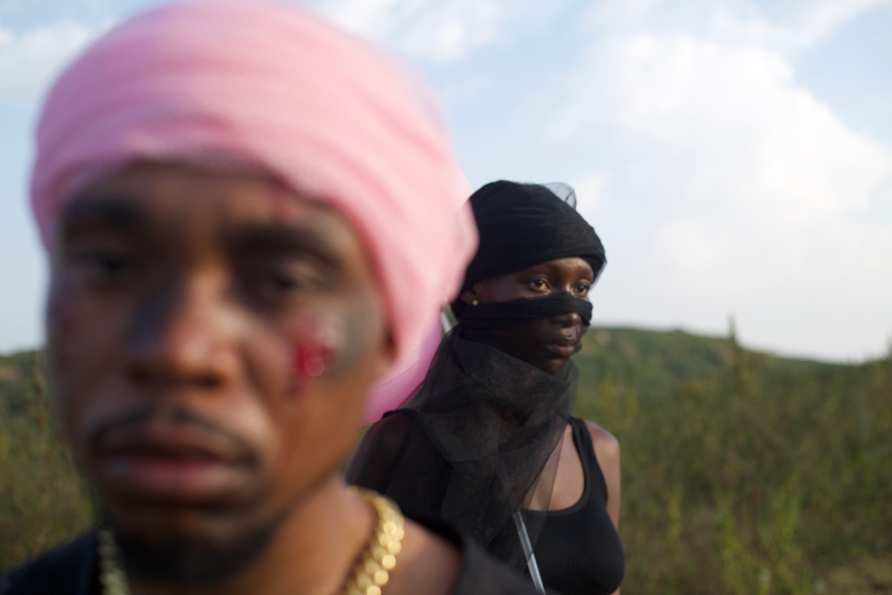 Two artists in a field at dusk: one in a pink headwrap in soft focus, another veiled in black tulle behind.